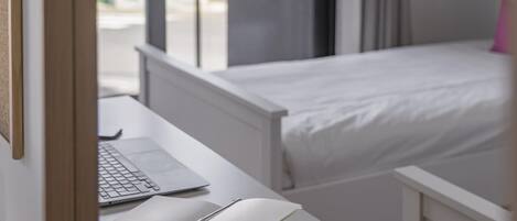 Un dormitorio con cama blanca, escritorio con computadora portátil y un libro abierto, y ventana con vista al exterior.