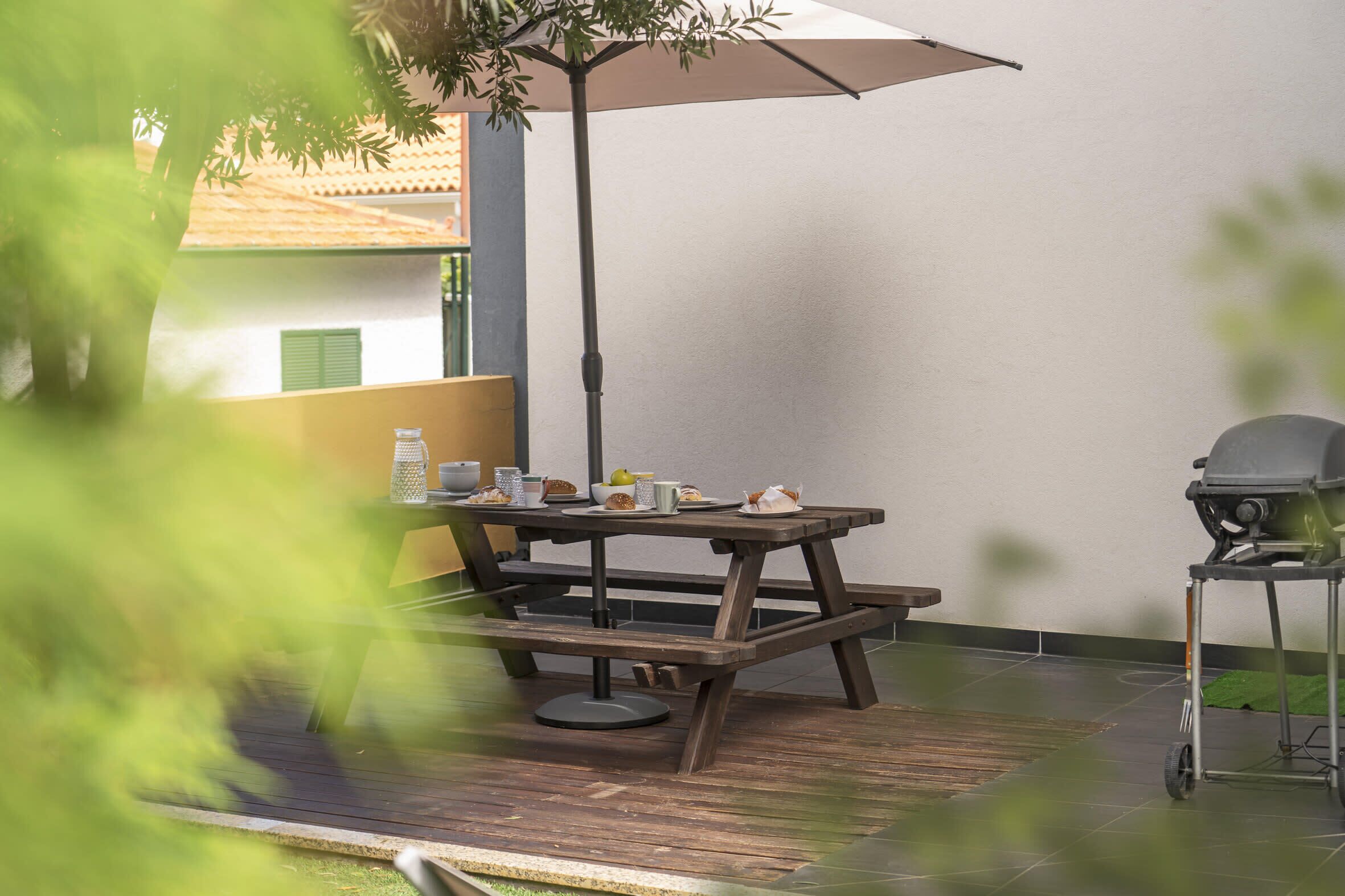 A wooden picnic table with a bench, a grill, and a table umbrella.