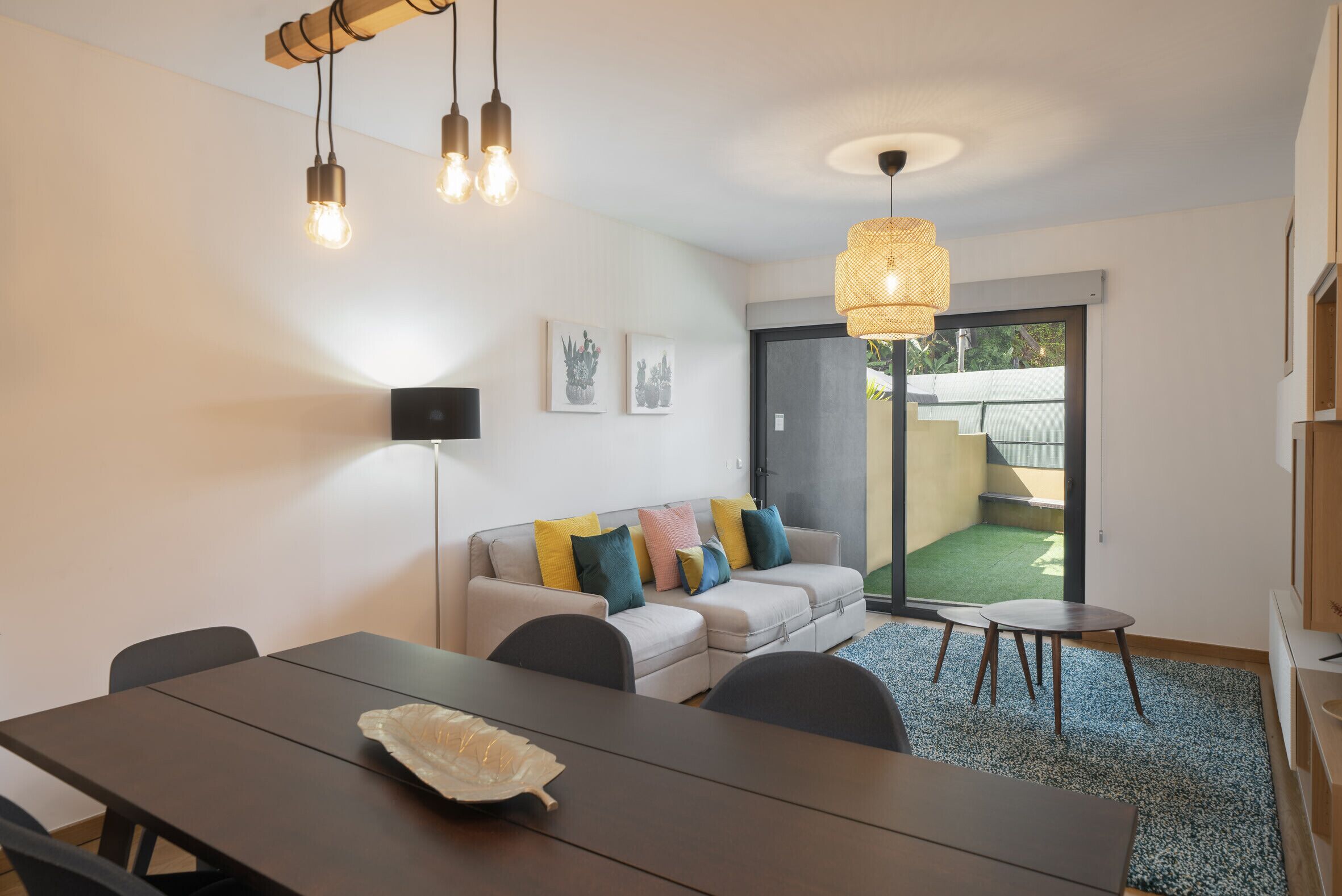A modern living room with a sofa, dining table, and chairs. There are wall-mounted lights and a ceiling light fixture.