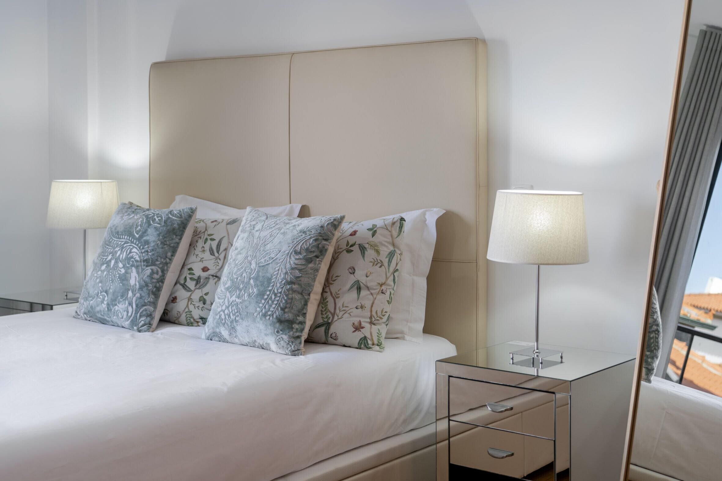 A neatly made bed with floral pillows, a bedside table with a lamp, and a view of the outside.