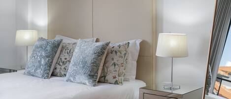 A neatly made bed with floral pillows, a bedside table with a lamp, and a view of the outside.