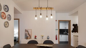 A modern dining area with a long table, chairs, a decorative light fixture, and a kitchen in the background. - In the Funchal Centre, with outdoor area - Pena II (Funchal)