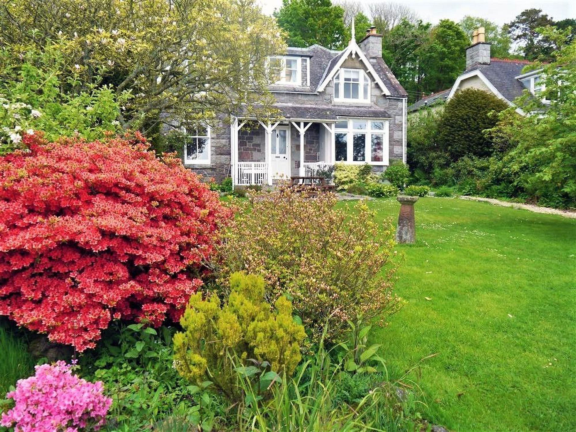 Une maison avec une façade en pierre, un porche et un jardin bien entretenu agrémenté de divers arbustes.