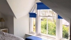 Un dormitorio con techo inclinado, una ventana con contraventanas azules y una cama con cabecera blanca.