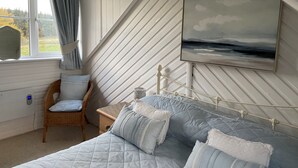 Un dormitorio con cama, una silla, una mesita de noche, un cuadro y una ventana con cortinas.