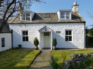 Une petite maison en pierre blanche avec une porte centrale, deux fenêtres à l’étage et une petite lucarne.
