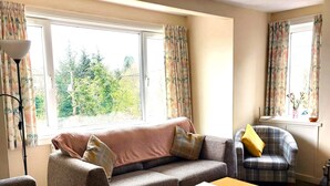 Una sala de estar con sofá, sillón y una mesa. Hay ventanas con cortinas, una lámpara y vista a follaje exterior.