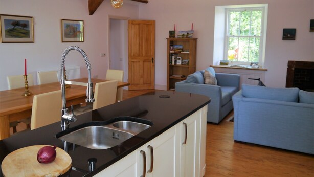 A kitchen with a double sink, wooden beams, a dining table with chairs, a blue sofa, and a bookshelf. - Rusko Stables -  an apartment that sleeps 6 guests  in 3 bedrooms (Gatehouse Of Fleet)