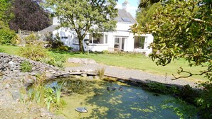 A pond with ducks, surrounded by a stone wall and greenery, with a house in the background.