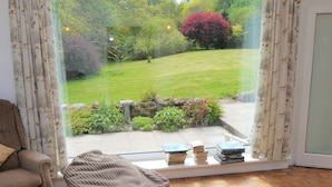 A room with a large window overlooking a garden, a sofa, a small table with books, and a wooden floor.