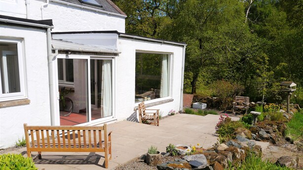 A white house with a patio, a wooden bench, and a garden with rocks and plants.