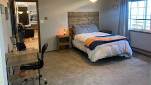 A bedroom with a bed, a desk with a chair, a ceiling fan, and a window.