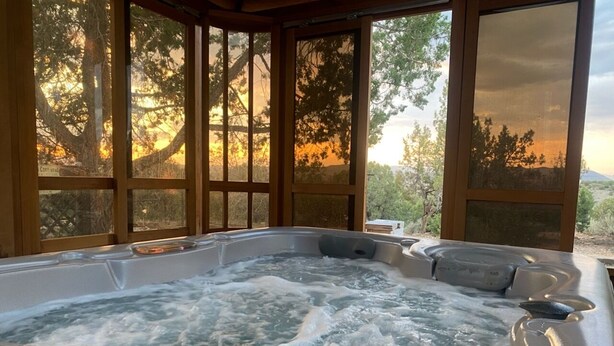 A hot tub with a view of trees and a sunset through large windows.