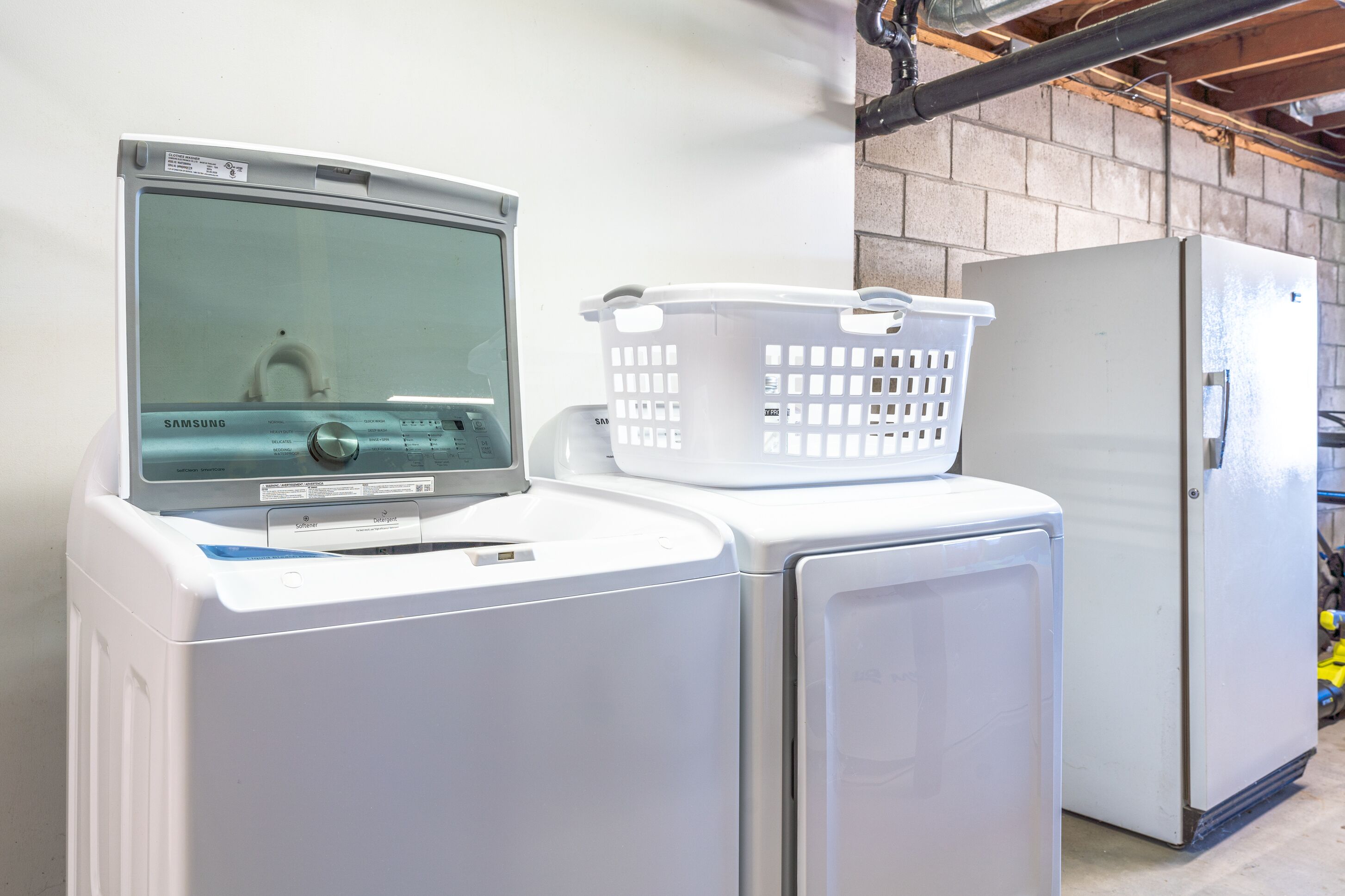 A front-loading washing machine with a control panel, a white plastic laundry basket on top, and a white refrigerator to the right.