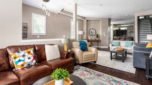 Een moderne woonkamer met een bruine leren bank, een beige fauteuil en een grijze bank met gele kussens. Er staat een bijzettafeltje met een plant op, een kamerkleed met dessin en er is een eethoek op de achtergrond.