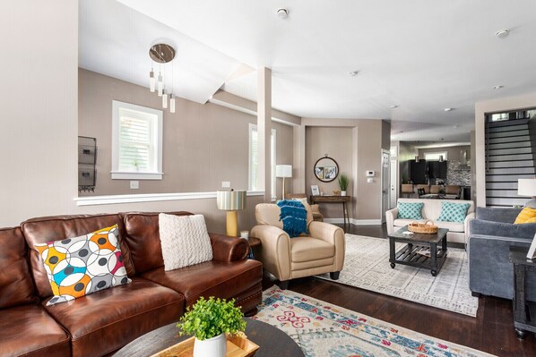 A modern living room with a brown leather sofa, beige armchair, and a grey sofa with yellow cushions. There is a coffee table with a plant, a patterned rug, and a dining area in the background.