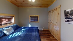 Een slaapkamer met een houten plafond, een bed met blauw beddengoed, een raam met zonwering, een radiator en een houten garderobe.