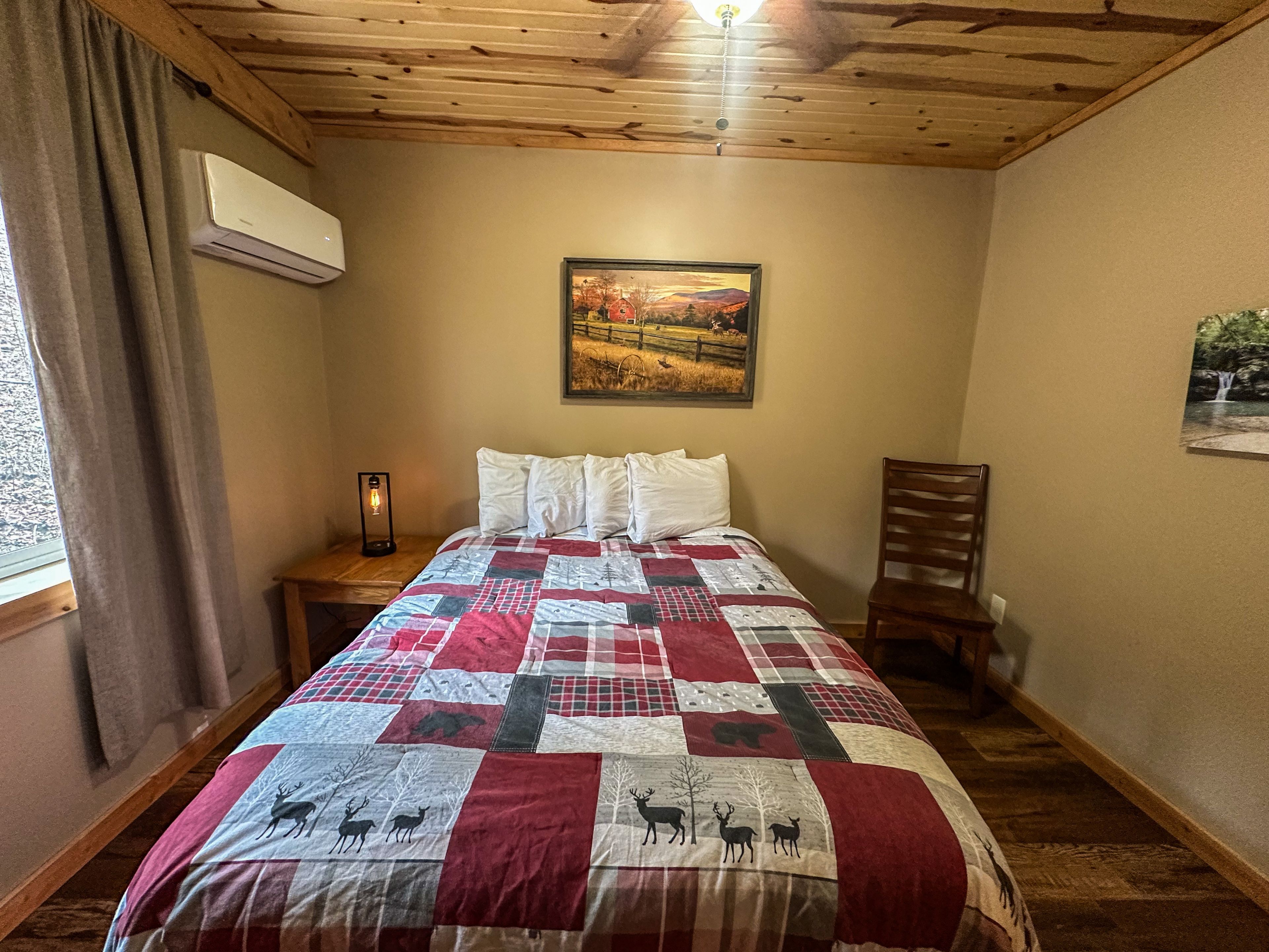 A bedroom with a wooden ceiling, a bed with a quilt featuring deer patterns, a nightstand with a lamp, and a painting of a landscape.