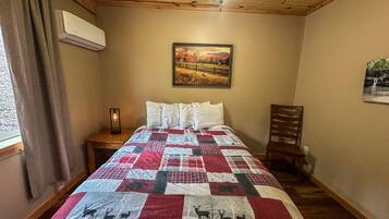 A bedroom with a wooden ceiling, a bed with a quilt featuring deer patterns, a nightstand with a lamp, and a painting of a landscape.