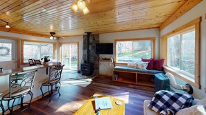 A cozy cabin interior with a stone fireplace, wooden floors, a dining area, and large windows offering views of the outdoors.