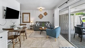 Una sala de estar moderna con un sofá, una mesa de centro y una televisión. Hay un comedor con sillas y una mesa. La habitación tiene un gran ventanal corredero que da a un espacio exterior.
