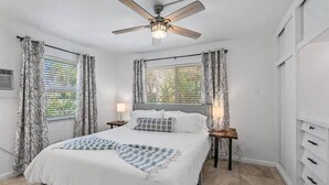 Un dormitorio con una cama, dos mesitas de noche con lámparas, un ventilador de techo y una ventana con cortinas.