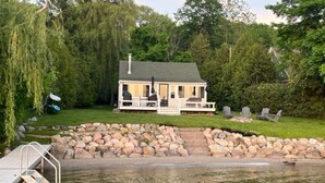 Una cabaña junto al lago con un muelle, rodeada por un muro de piedra y abundante vegetación.