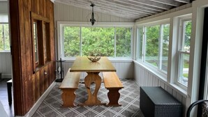 Un porche cubierto con una mesa de madera y bancos, ventanas amplias y vistas a la vegetación.