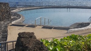 Una piscina con pared de piedra y barandilla metálica, rodeada por una zona arenosa y vegetación.