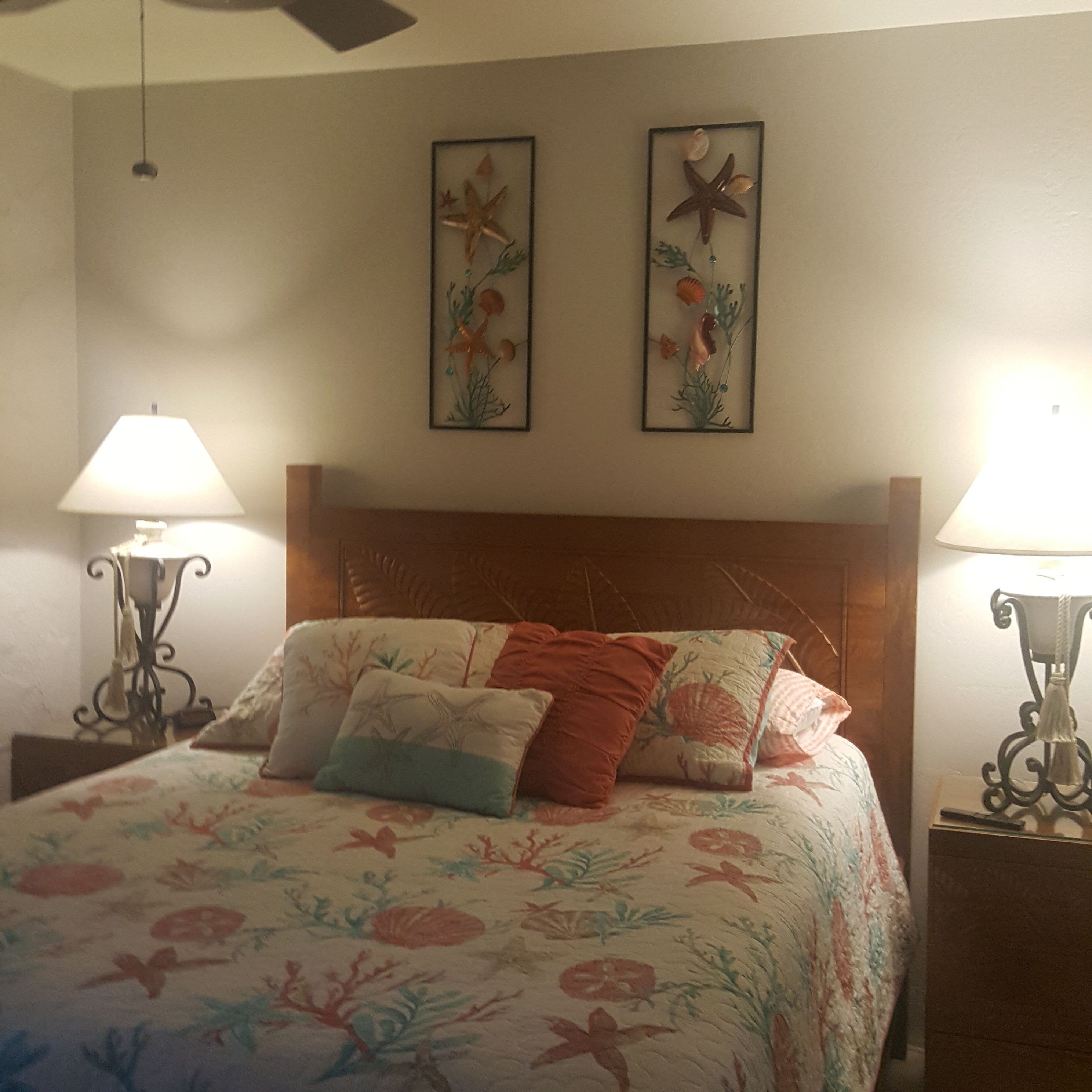 Quarto com cabeceira de madeira, roupa de cama florida, duas estrelas-do-mar decorativas na parede e duas luminárias de cabeceira.