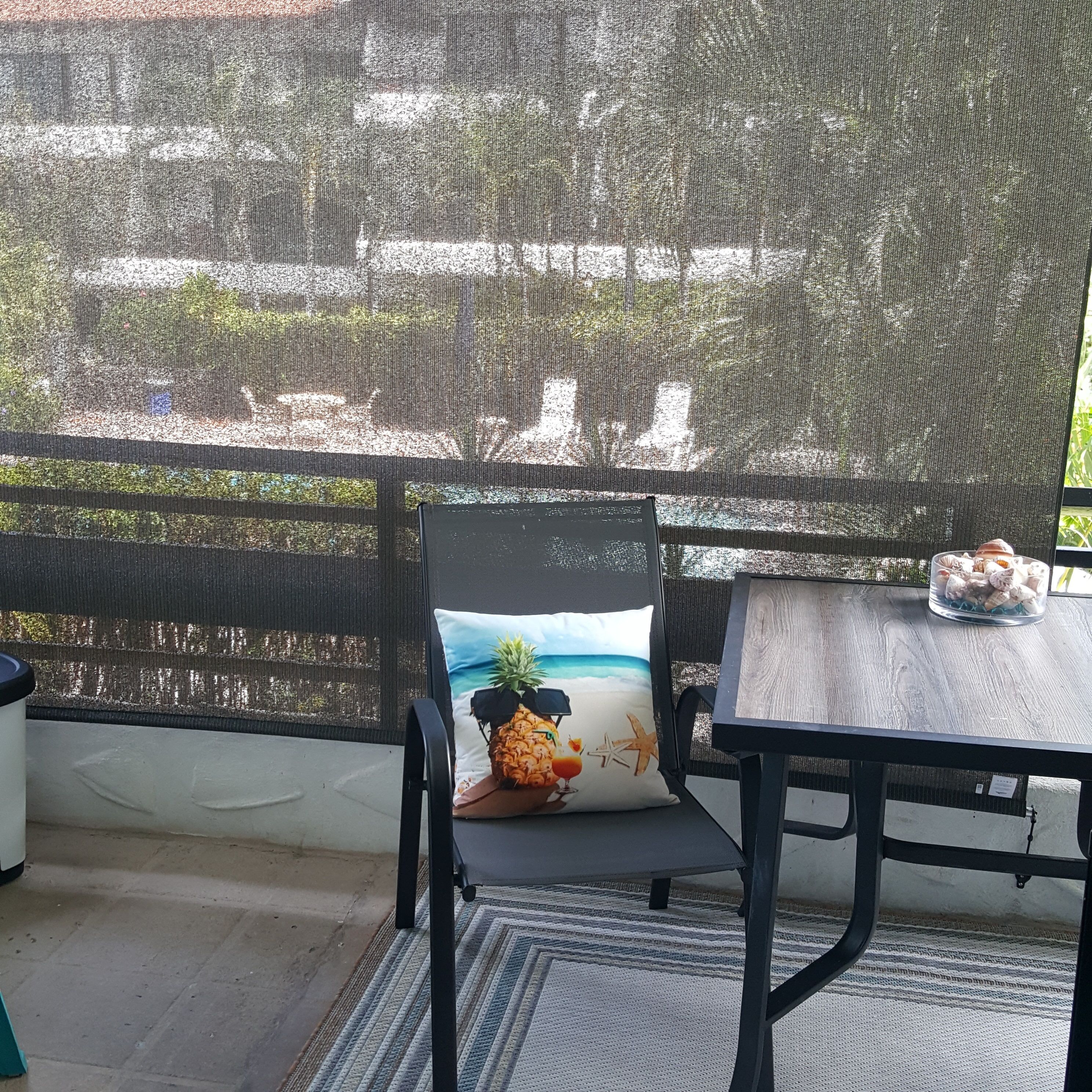 Un balcón con mosquitera, una silla con cojín de motivos tropicales, una mesita con un bol de aperitivos y vistas a un edificio y follaje.