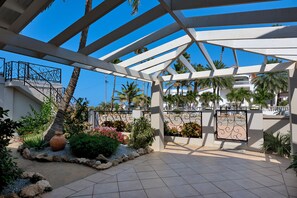 Een overdekt buitengebied met een witte pergola, palmbomen en uitzicht op een gebouw met een rood dak.