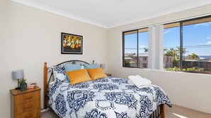 Ein Schlafzimmer mit Bett, Nachttisch, Lampe, einem gerahmten Bild und einem Fenster mit Blick auf Gebäude und Palmen.