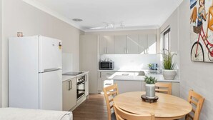 Eine kompakte Küche mit weißen Geräten, ein Essbereich mit Holztisch und -stühlen sowie ein Topf mit Pflanze auf der Arbeitsfläche.