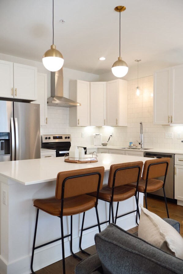 A modern kitchen with white cabinetry, a stainless steel refrigerator, a built-in oven, and a central island with bar stools.