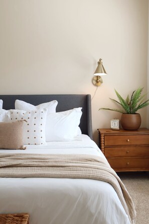 A neatly made bed with white bedding and multiple pillows, a bedside table with a clock, a potted plant, and a wall-mounted lamp.