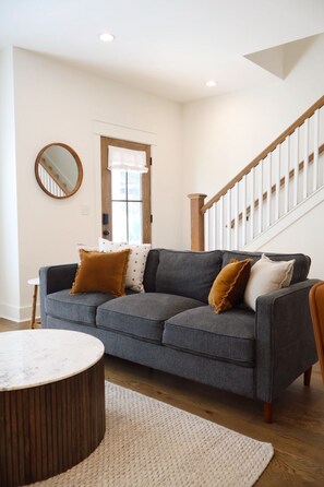 A living room with a gray sofa, a round wooden coffee table, a white rug, a wooden staircase, and a mirror on the wall.