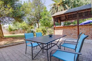 Een terras buiten met een bakstenen muur, een houten pergola en een tafel met blauwe stoelen.