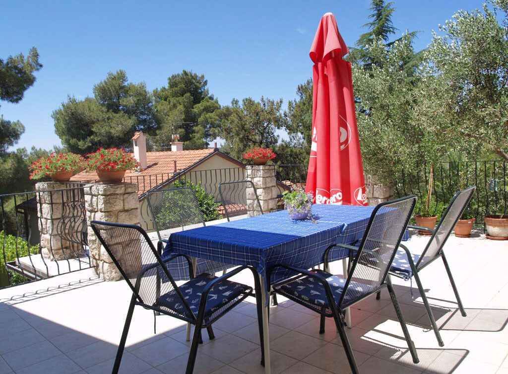 Une terrasse extérieure avec une table bleue et quatre chaises, un parasol rouge et des plantes en pot.