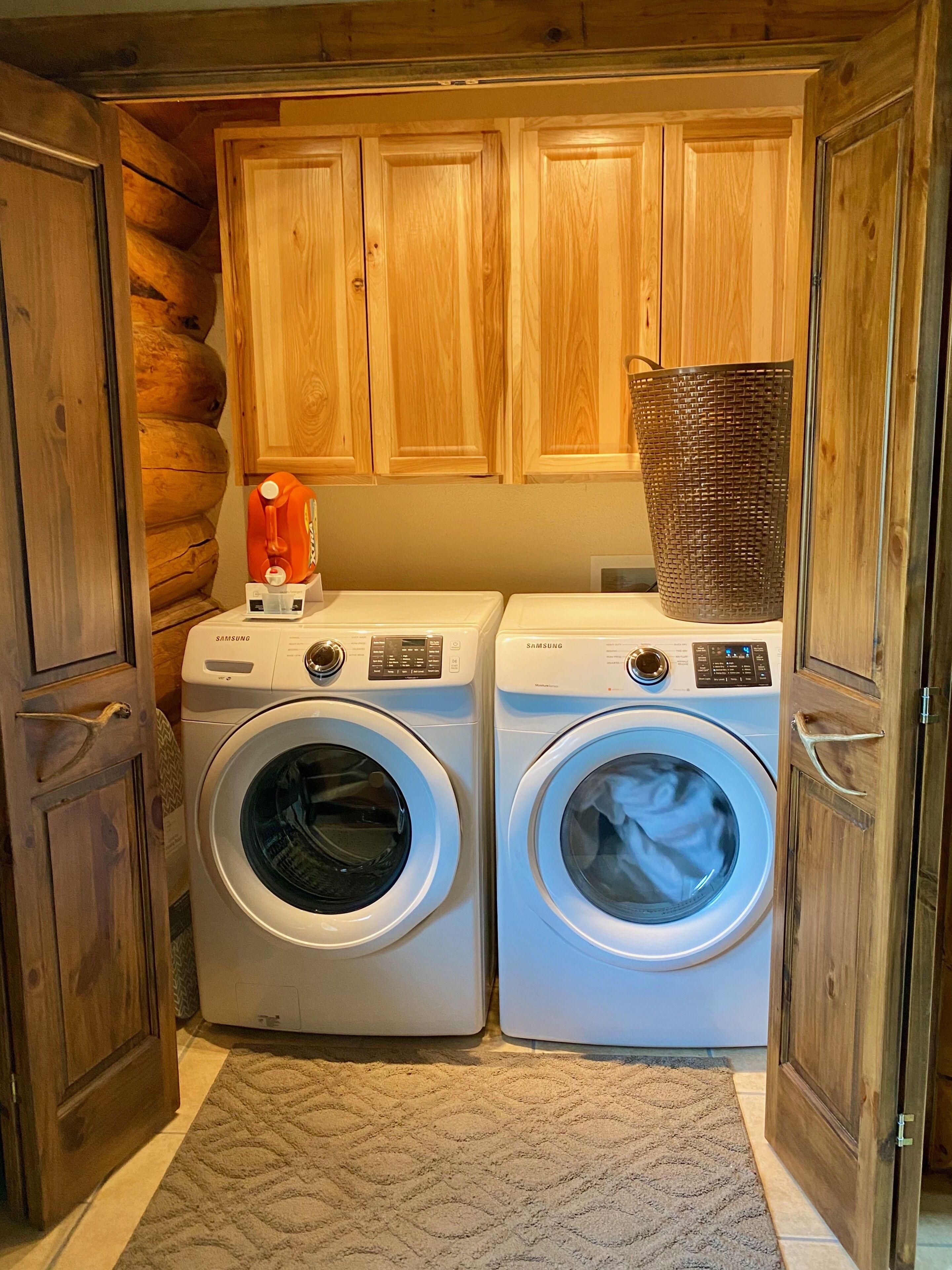 A laundry room with two Samsung washing machines, wooden cabinets, and a basket on top of one machine.