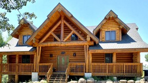 Une cabane en rondins avec un porche à l’avant, une porte centrale et deux fenêtres de chaque côté.