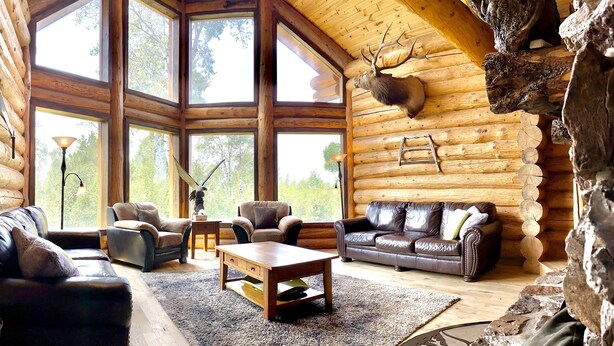 L'intérieur d'une cabane en rondins, avec une grande fenêtre, des canapés en cuir, une table basse en bois et une tête de cerf fixée au mur.