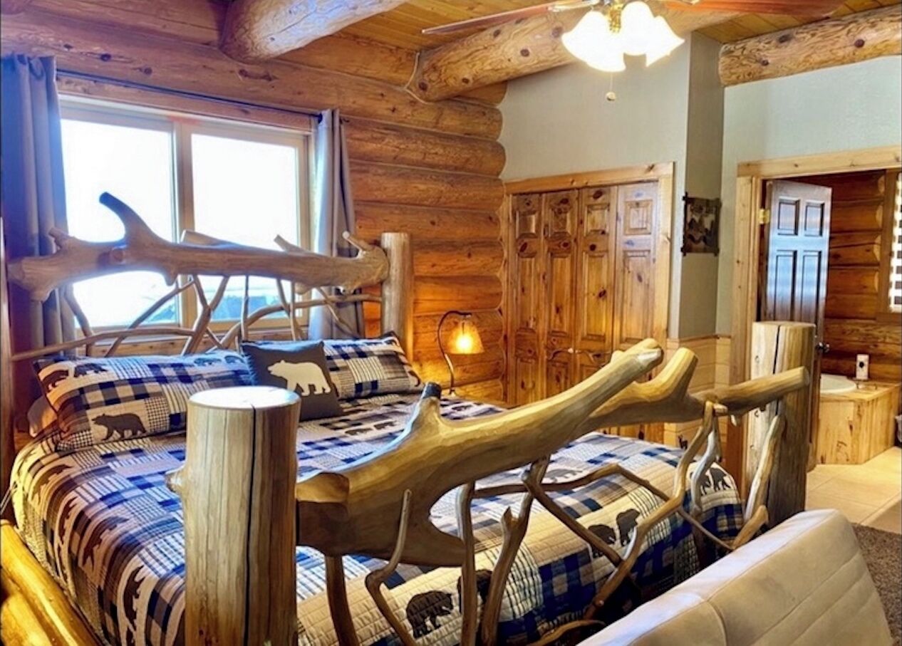 Interior de una cabaña de troncos con una cama que tiene reposacabezas de asta de ciervo, un armario de madera y una ventana con cortinas.