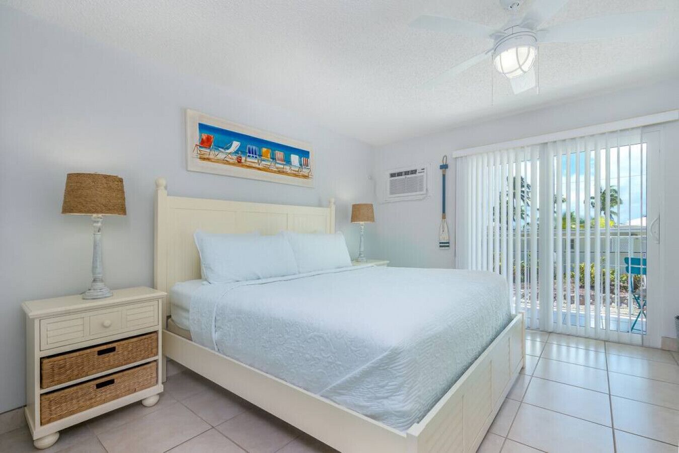 A bedroom with a bed, two bedside tables, a ceiling fan, a window with blinds, and a painting on the wall.