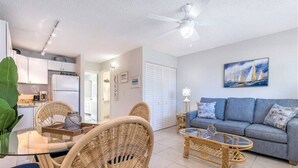 Ein modernes Wohnzimmer mit einer Couch, einem Couchtisch und Korbstühlen. Es gibt einen Deckenventilator, ein Gemälde an der Wand und eine Küchenzeile im Hintergrund.