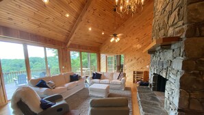 A cozy cabin interior with a stone fireplace, wooden ceiling, and large windows offering a view of the forest. - Views, hot tub, spacious 3Bd, 3Ba w Tesla Charger (Robbinsville)