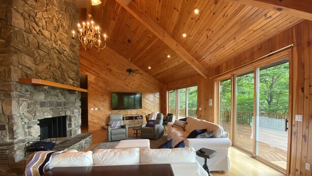 A cozy cabin interior with a stone fireplace, wooden ceiling, and large windows offering a view of the outdoors. - Views, hot tub, spacious 3Bd, 3Ba w Tesla Charger (Robbinsville)