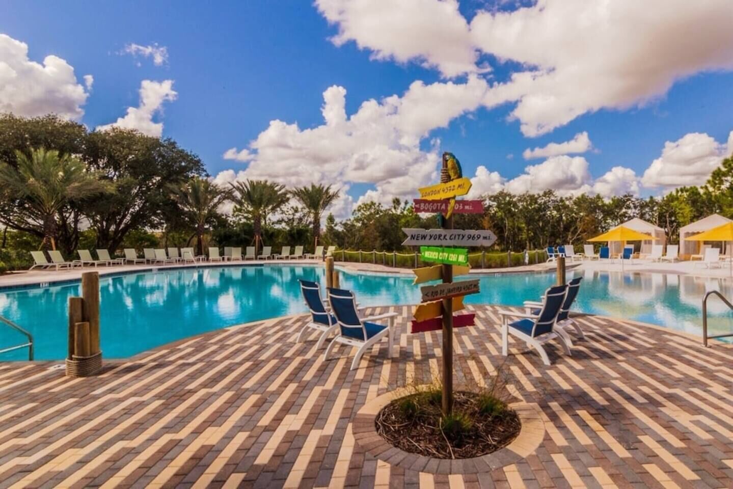 Un espace aménagé au bord de la piscine, avec des chaises longues, un panneau indicateur portant les noms de différentes villes, et un ciel d’un bleu limpide.