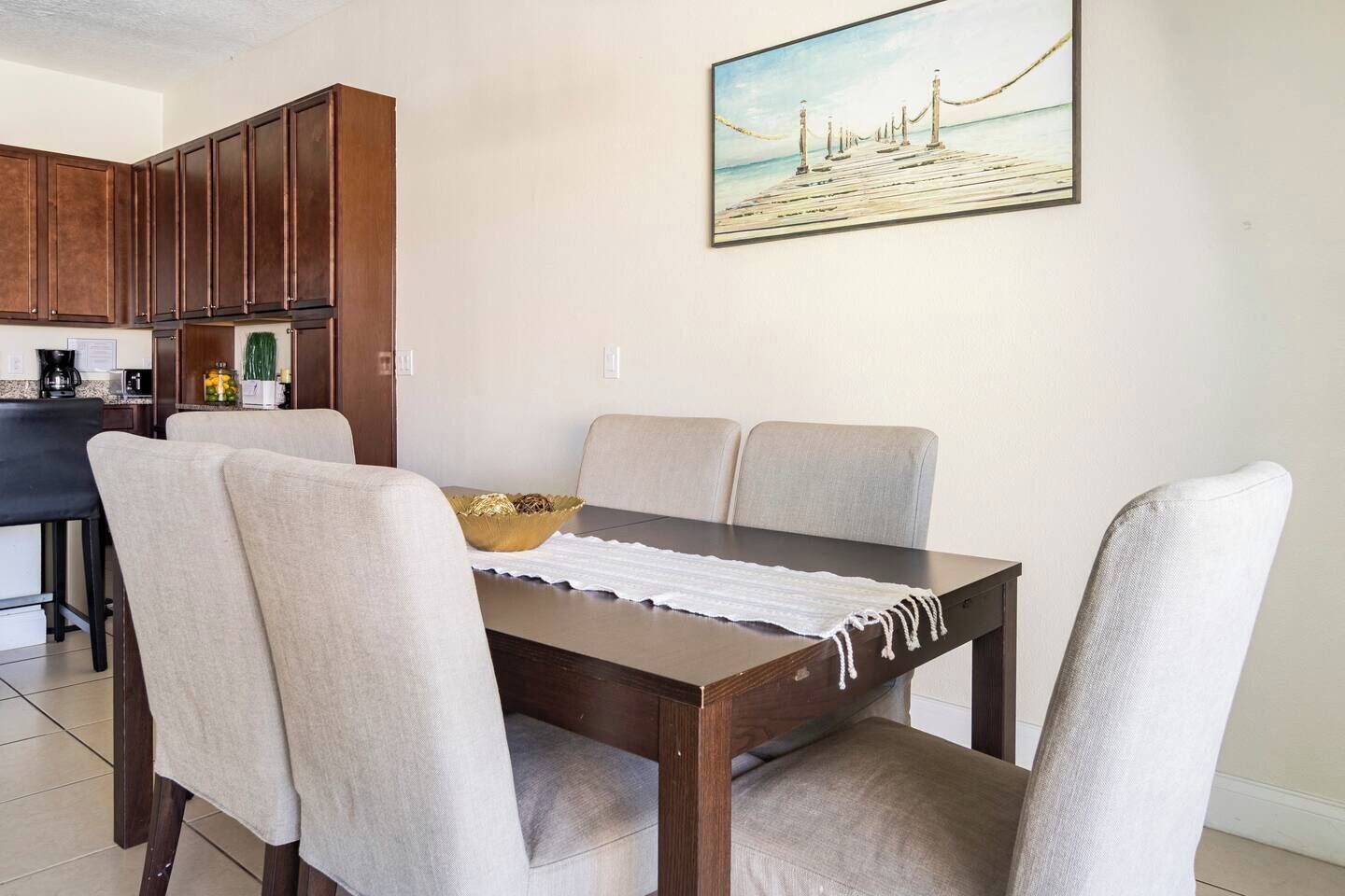 Un comedor con una mesa de madera, sillas color beige y un cuadro de una playa.