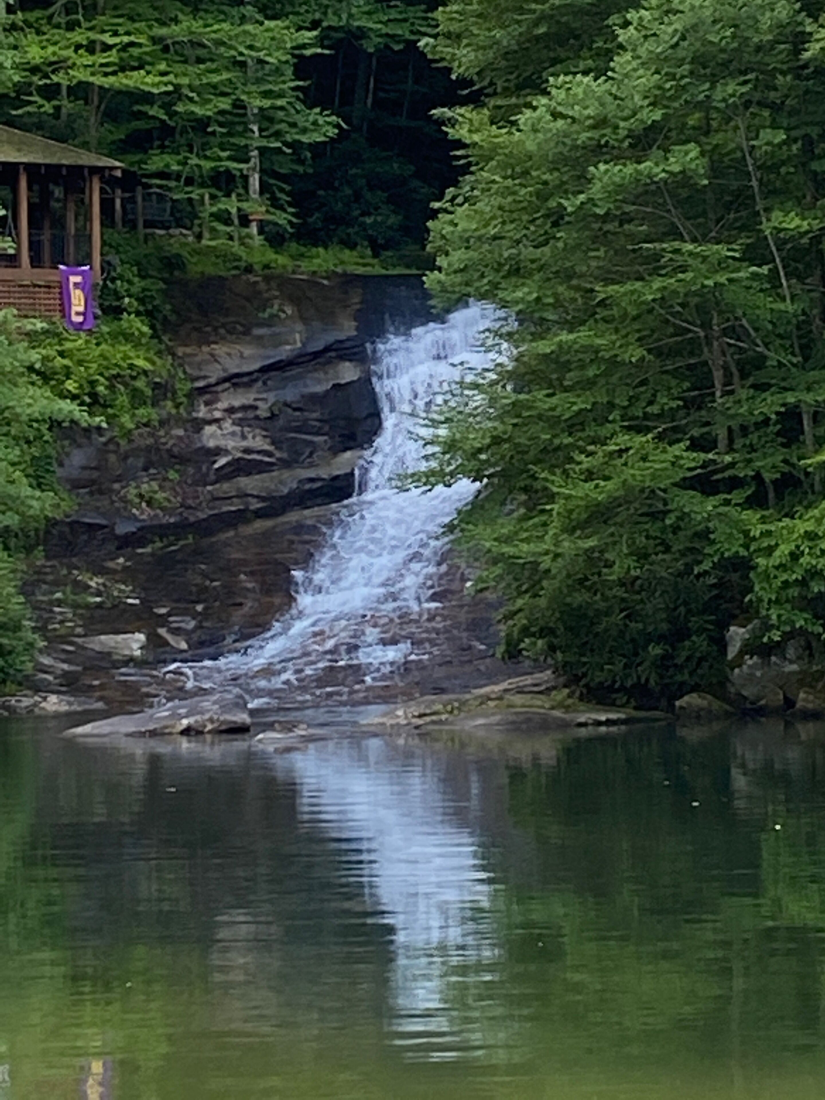 A waterfall cascading into a calm body of water, surrounded by dense green foliage and a wooden structure with a purple sign.